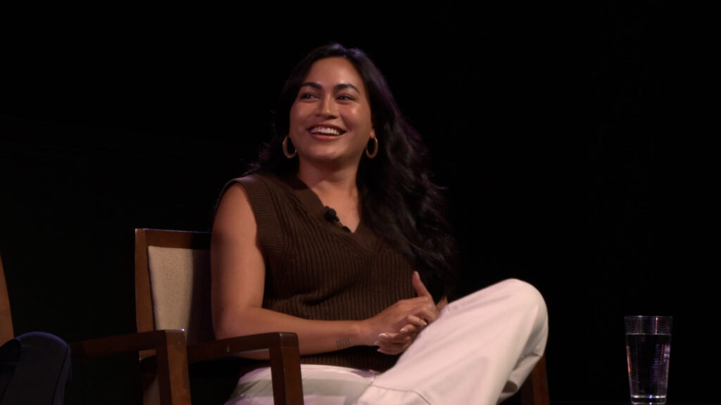 Lindsay Watson, actor, The Wind and the Reckoning, sits for a post-screening discussion. She wears a dark brown sleeveless sweater, beige pants and thick golden hoops for earrings. He hands are clasped and she is smiling towards the left of the screen.