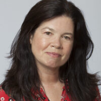 Filmmaker Pamela Yates appears against a light grey backdrop. She is facing the camera and smiling slightly. She has long brown hair and is wearing a red flowered blouse.