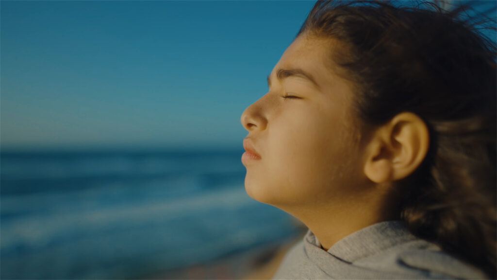 A still from the film From Ground Zero. It shows a girl standing on the beach, eyes closed, wind in her hair, taking a deep breathe in.